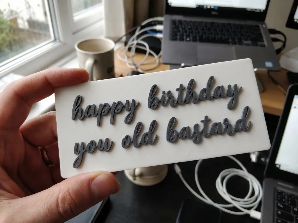 A 3D-printed Thudletter slab with raised handwritten lettering reading 'happy birthday you old bastard', shown in a real home setting.
