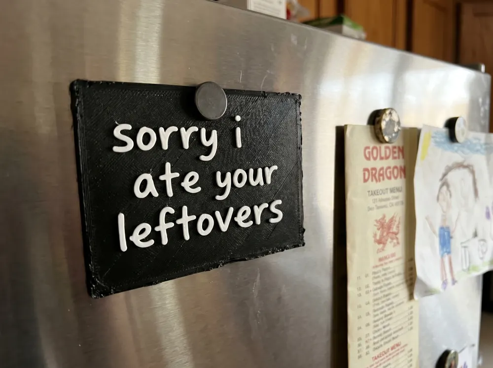 A 3D-printed Thudletter slab with raised handwritten lettering reading 'sorry i ate your leftovers', shown in a real home setting.
