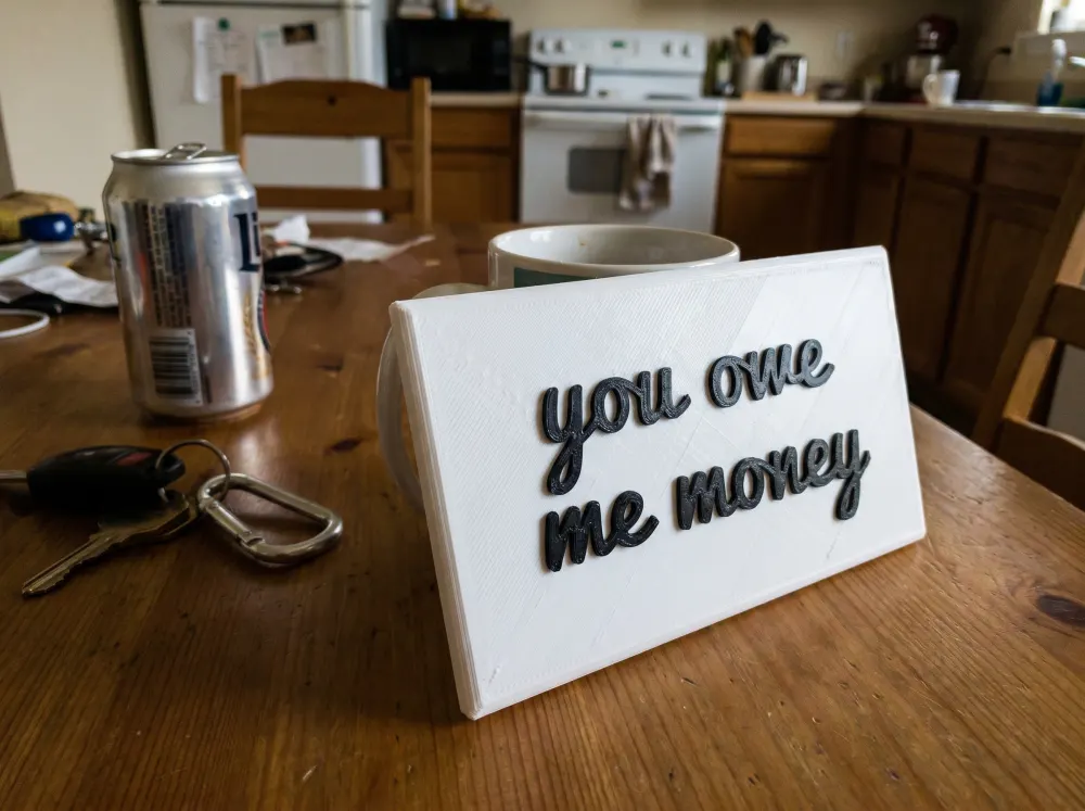 A 3D-printed Thudletter slab with raised handwritten lettering reading 'you owe me money', shown in a real home setting.
