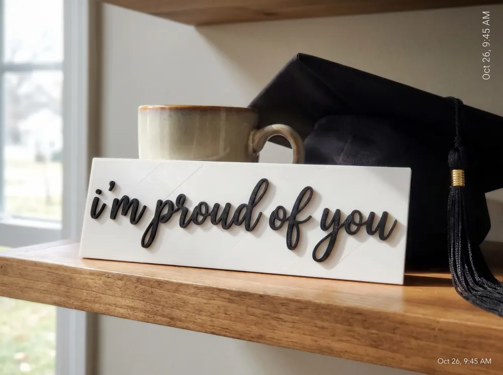 A 3D-printed Thudletter slab with raised handwritten lettering reading 'i'm proud of you', shown in a real home setting.