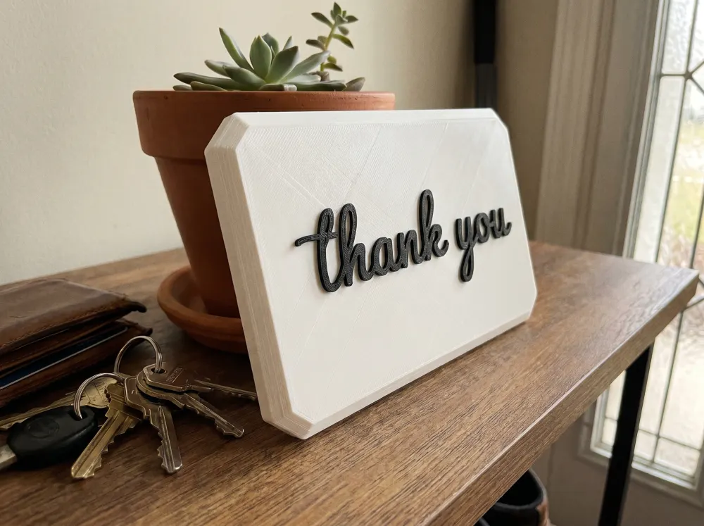A 3D-printed Thudletter slab with raised handwritten lettering reading 'thank you', shown in a real home setting.